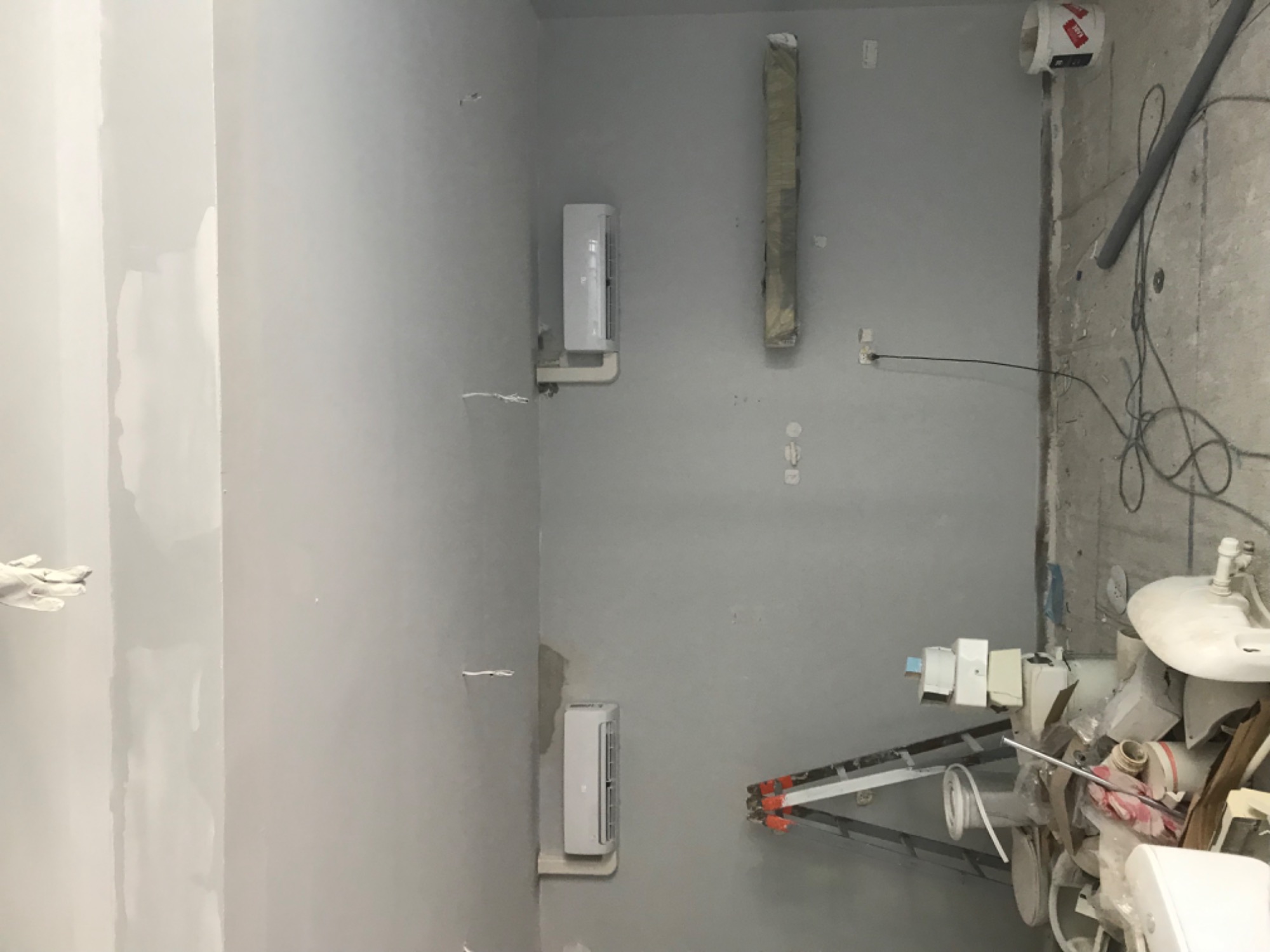 Wnętrze w trakcie remontu z dwoma zamontowanymi jednostkami klimatyzacyjnymi na szarej ścianie, widoczne przewody, drabina i pozostałości materiałów budowlanych.