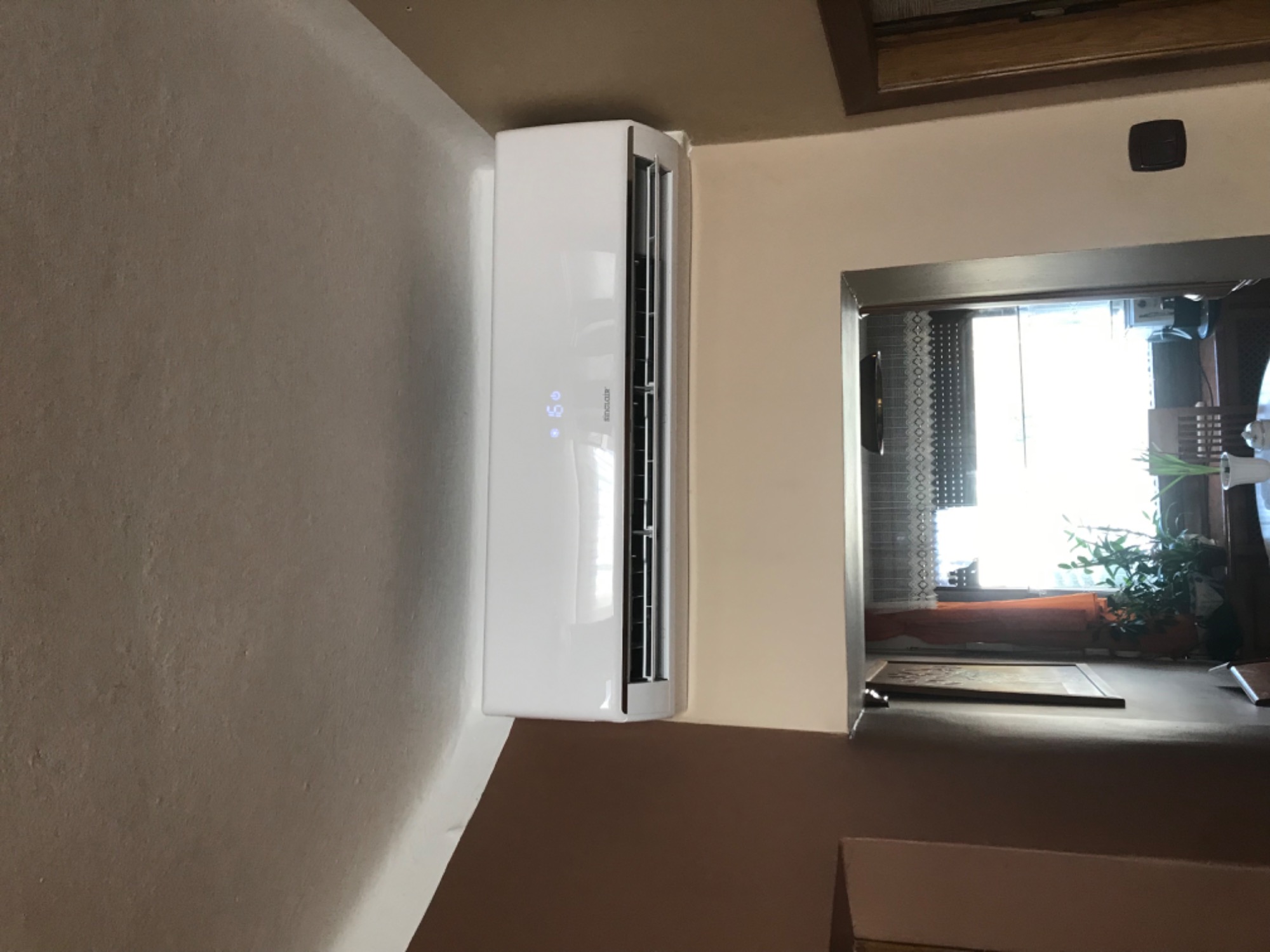 Biała klimatyzacja Sinclair zamontowana na ścianie w pomieszczeniu z widokiem na okno z roślinami.