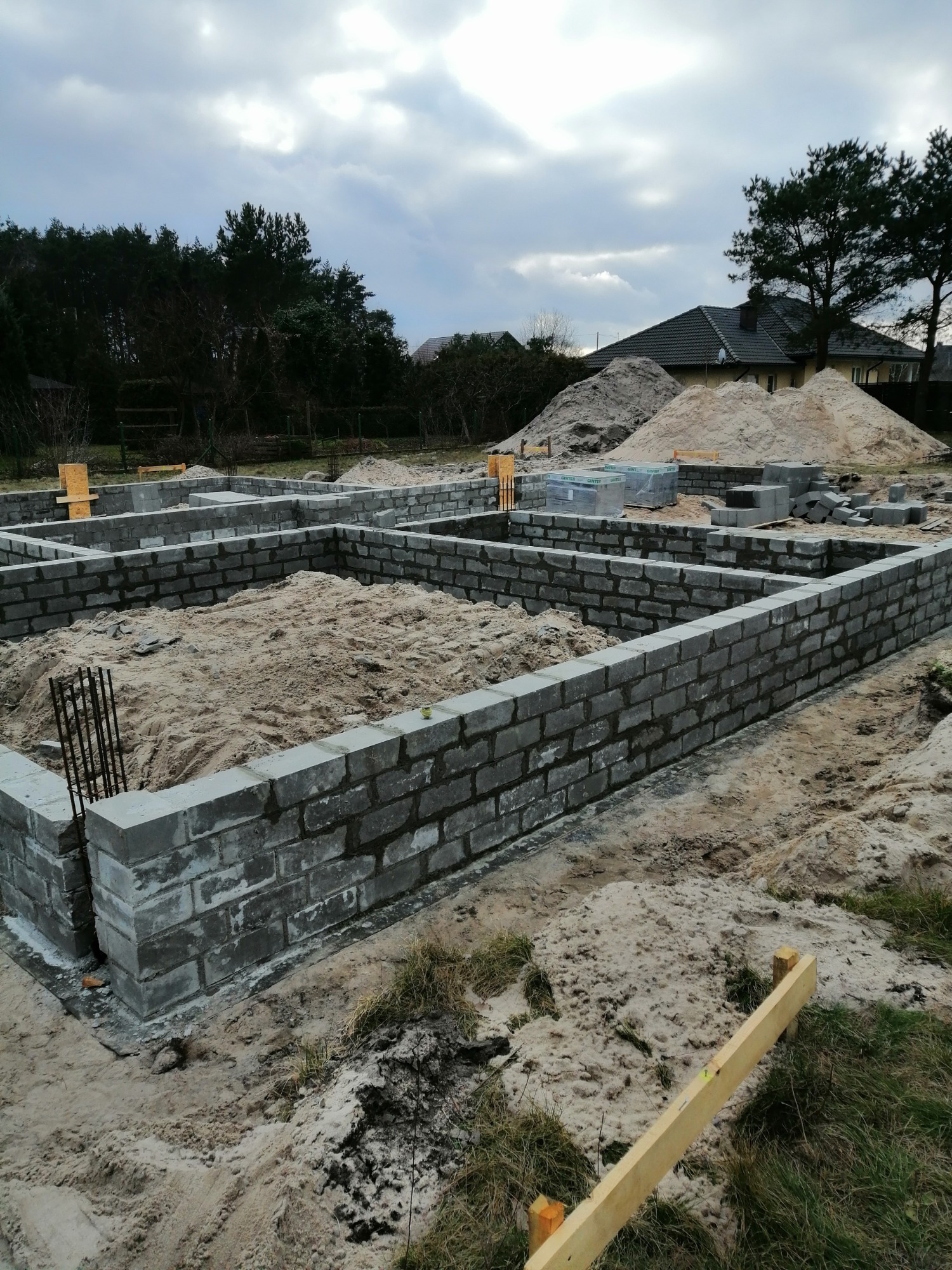 Fundamenty z bloczków betonowych wypełnione piaskiem, widoczne zbrojenia, w tle hałda piasku i dom w budowie, pochmurne niebo.