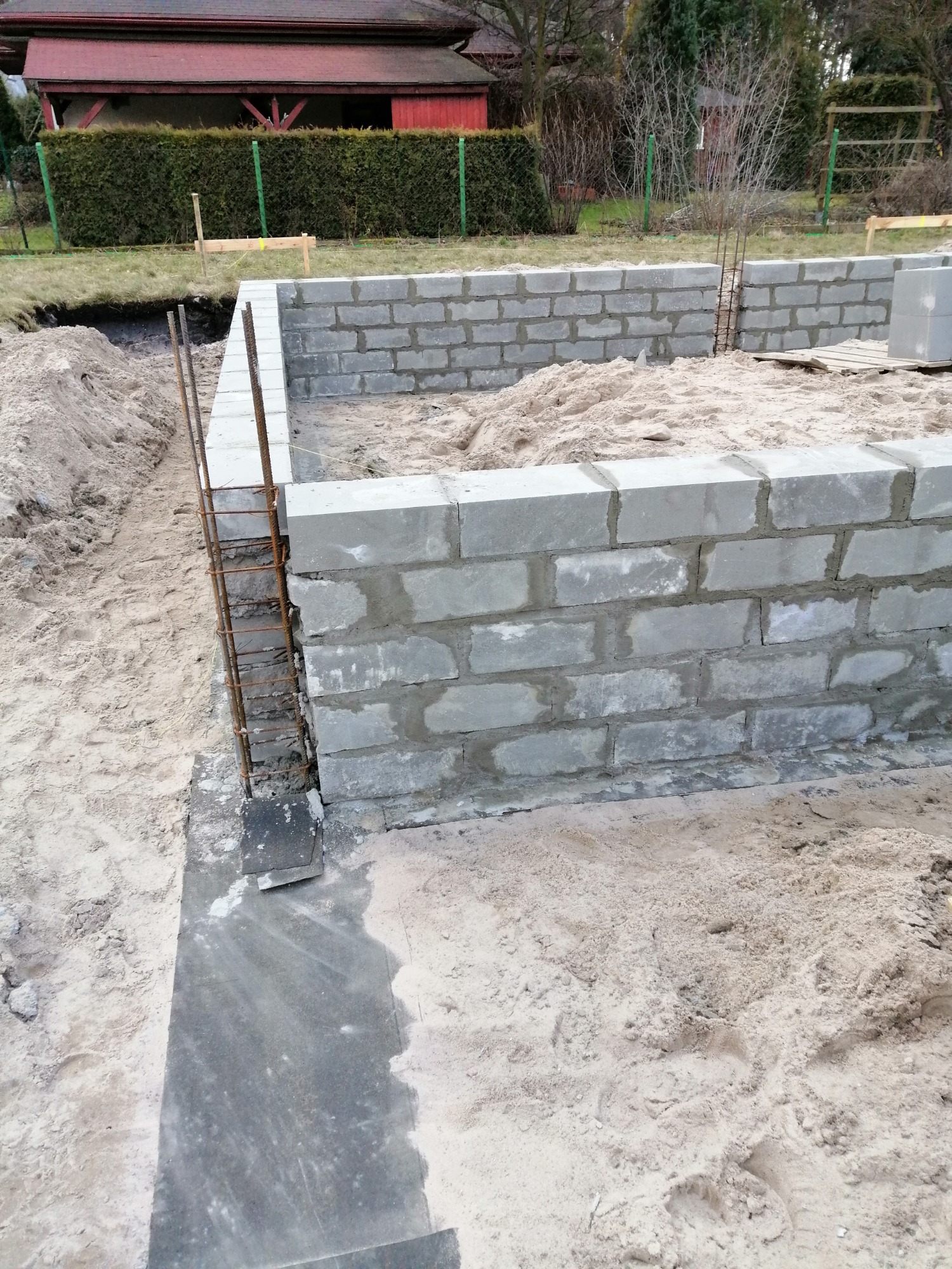 Wznoszenie fundamentów z bloczków betonowych z wystającymi prętami zbrojeniowymi, widoczny piasek wewnątrz i na zewnątrz fundamentów, w tle budynek mieszkalny.