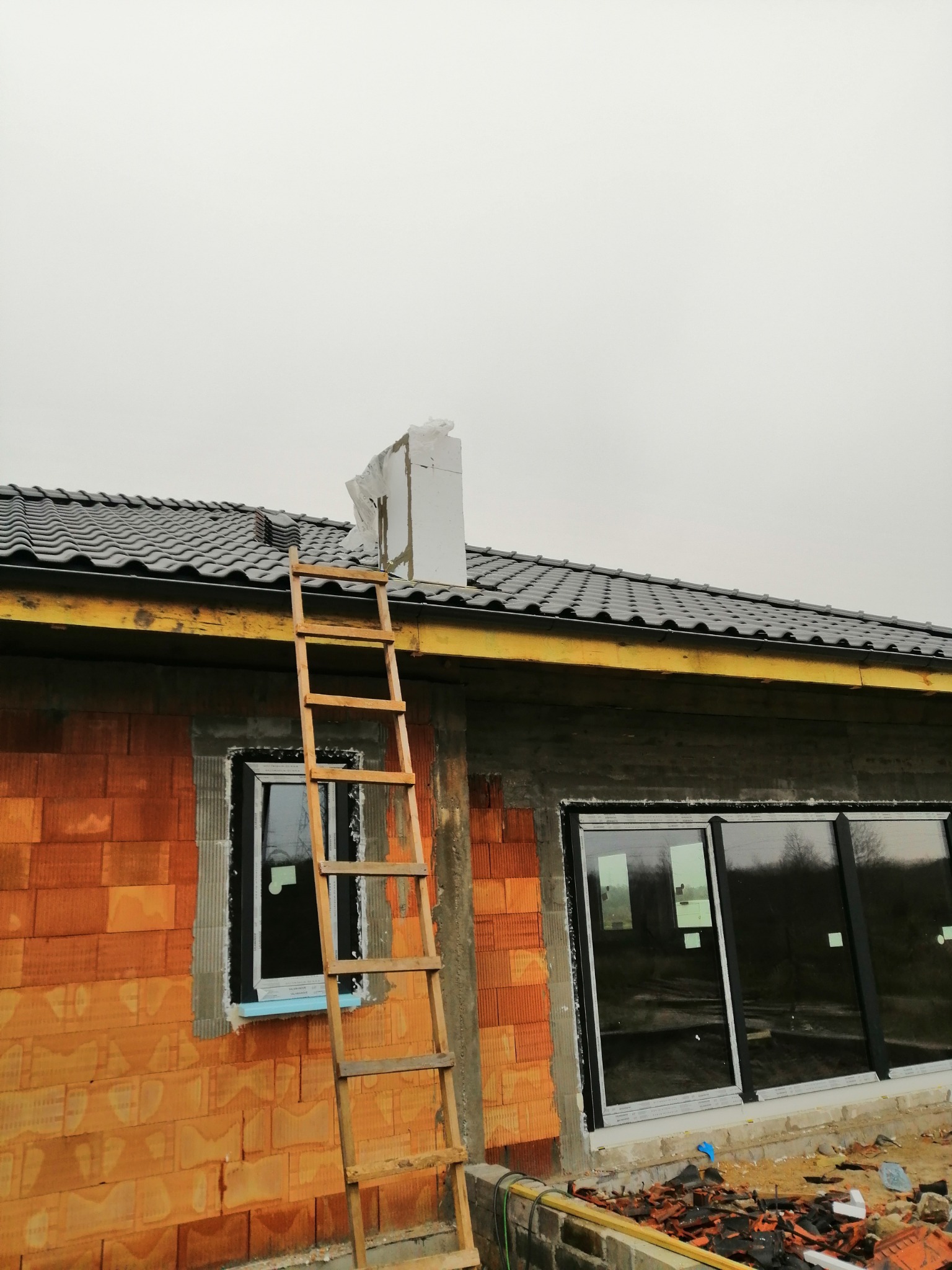 Budowa domu w trakcie realizacji: widoczna murowana ściana z oknami, drewniana drabina oparta o ścianę, fragment dachu z ciemną dachówką i komin zabezpieczony folią.