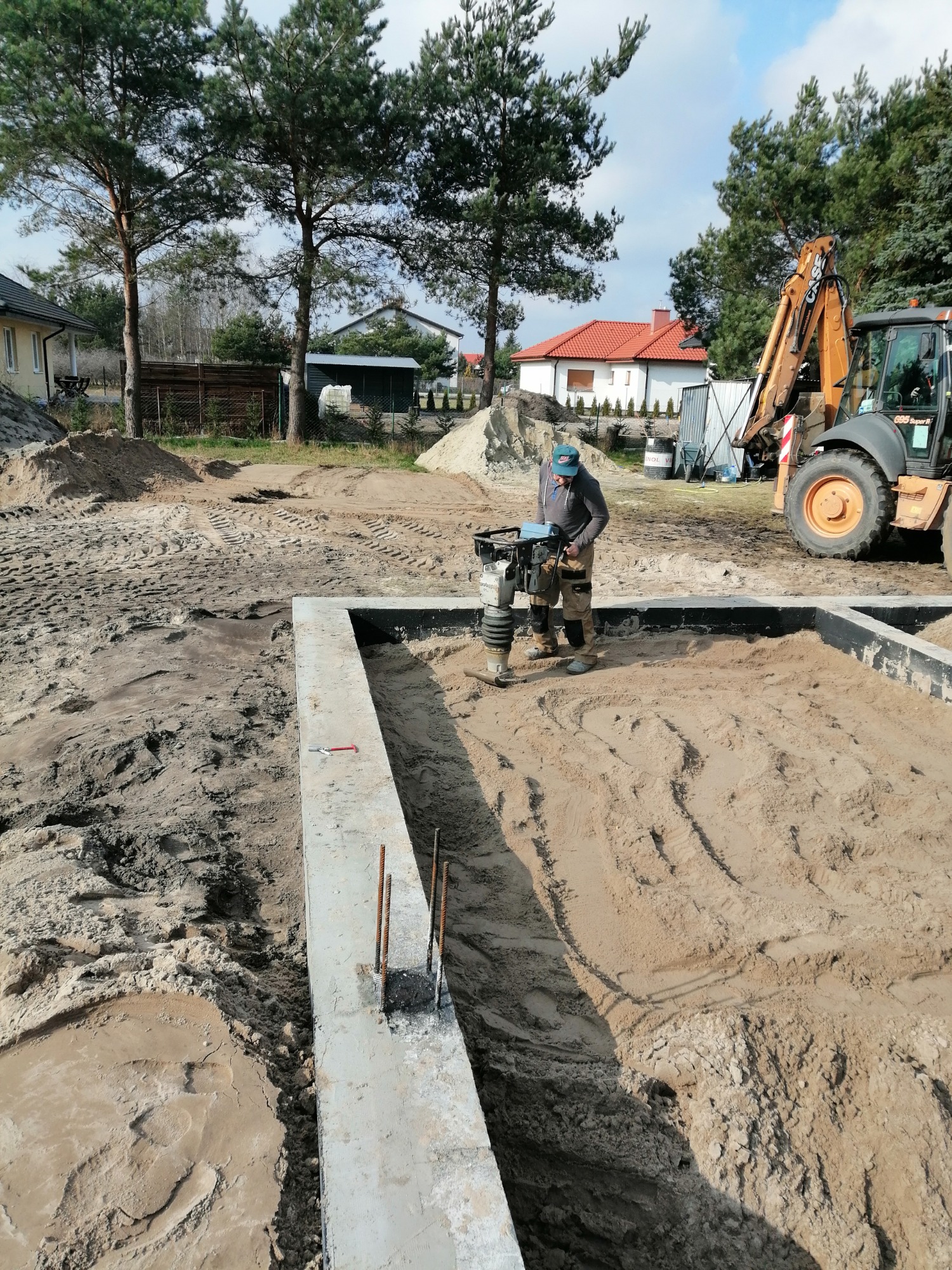 Pracownik zagęszcza piasek w fundamencie domu jednorodzinnego za pomocą zagęszczarki wibracyjnej, widoczny szalunek i wystające pręty zbrojeniowe, w tle koparka i zabudowa jednorodzinna.