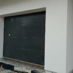 Nowoczesna, segmentowa brama garażowa w kolorze antracytowym, z widoczną instalacją drenażową u podstawy ściany i fragmentem drzwi wejściowych z elementami stali nierdzewnej.