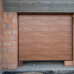 Zamknięta brama garażowa segmentowa z imitacją drewna, widoczna w surowym otoczeniu budowy, z niedokończonymi ścianami z cegieł i betonowym nadprożem.