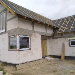 Niedokończony budynek z białych bloczków z zamontowanymi oknami z brązowymi ramami i dachem w trakcie budowy, widoczne drewniane łaty i papa.