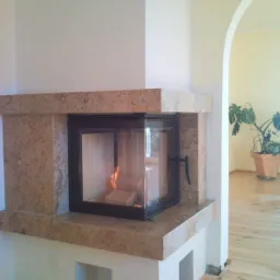 Nowoczesny kominek z obudową z jasnego granitu, z paleniskiem zamkniętym czarną ramą i widocznym płomieniem, umieszczony w rogu jasnego pokoju z drewnianą podłogą i łukowatym przejściem do dalszej...