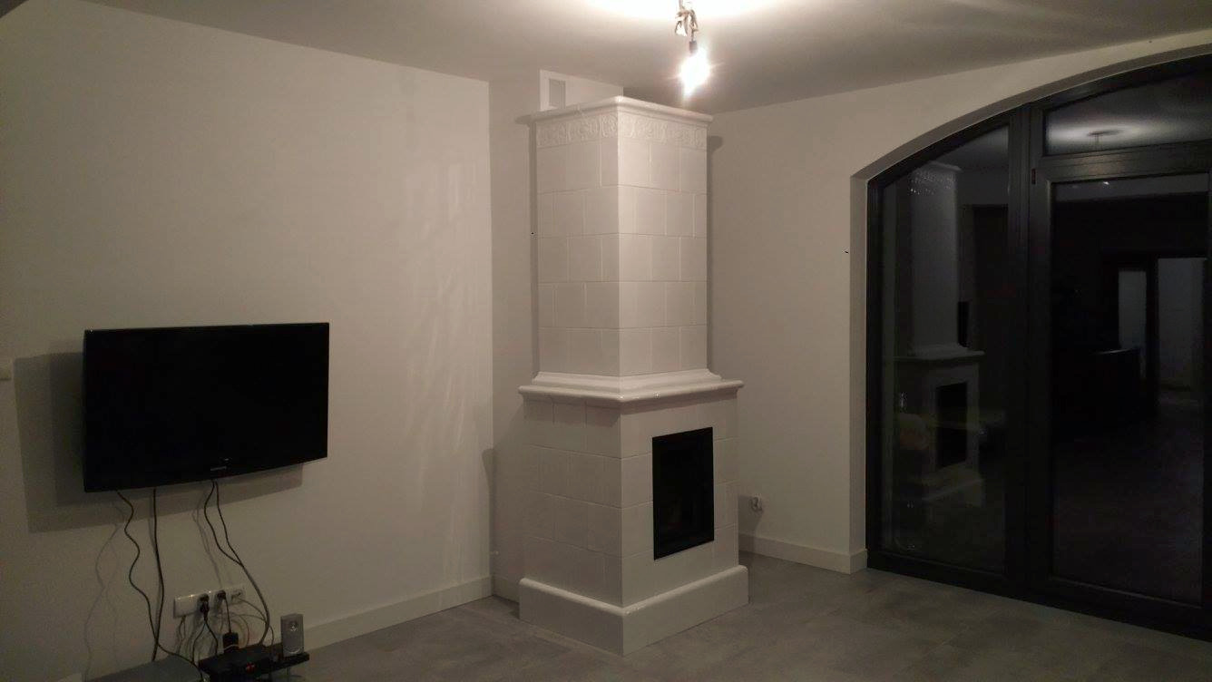 Biały, kaflowy piec kominkowy w nowoczesnym salonie z szarymi płytkami podłogowymi, obok telewizor na ścianie i przeszklone drzwi balkonowe.