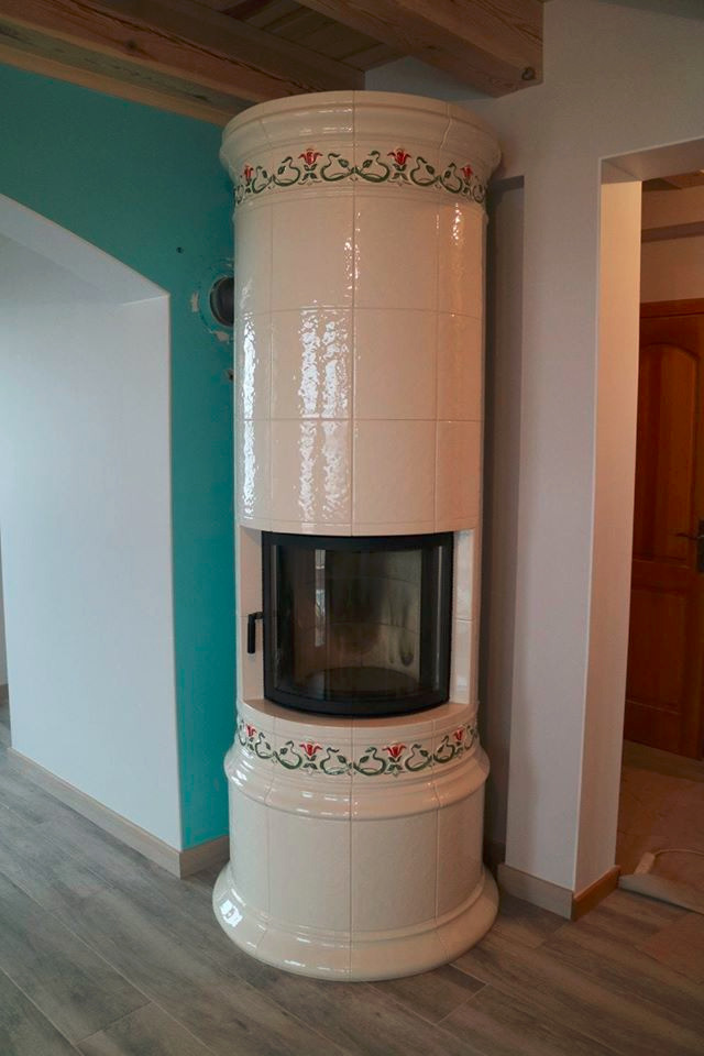 Stojący piec kaflowy w kolorze kremowym z dekoracyjnym motywem kwiatowym, umieszczony w rogu pomieszczenia z drewnianą podłogą i turkusową ścianą z widocznym otworem wentylacyjnym.