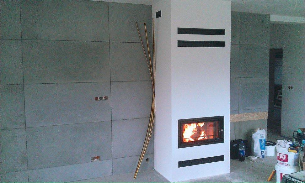 Nowoczesny kominek z czarną ramą, płomieniami wewnątrz, dekoracyjnymi złotymi prętami i szarą ścianą z płyt, w trakcie wykańczania.