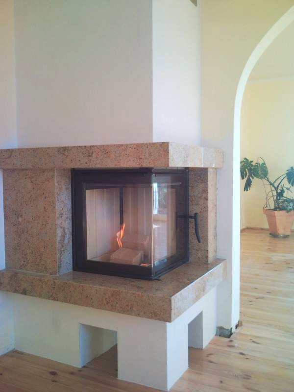 Nowoczesny kominek z obudową z jasnego granitu, z paleniskiem zamkniętym czarną ramą i widocznym płomieniem, umieszczony w rogu jasnego pokoju z drewnianą podłogą i łukowatym przejściem do dalszej...