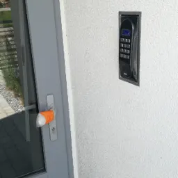 Zewnętrzny panel domofonu z klawiaturą numeryczną zamontowany na białej, teksturowanej ścianie obok drzwi z zabezpieczoną klamką.