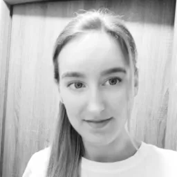 Czarno-białe portretowe zdjęcie młodej kobiety z włosami związanymi w kucyk, uśmiechającej się delikatnie do kamery na tle drewnianej ściany.