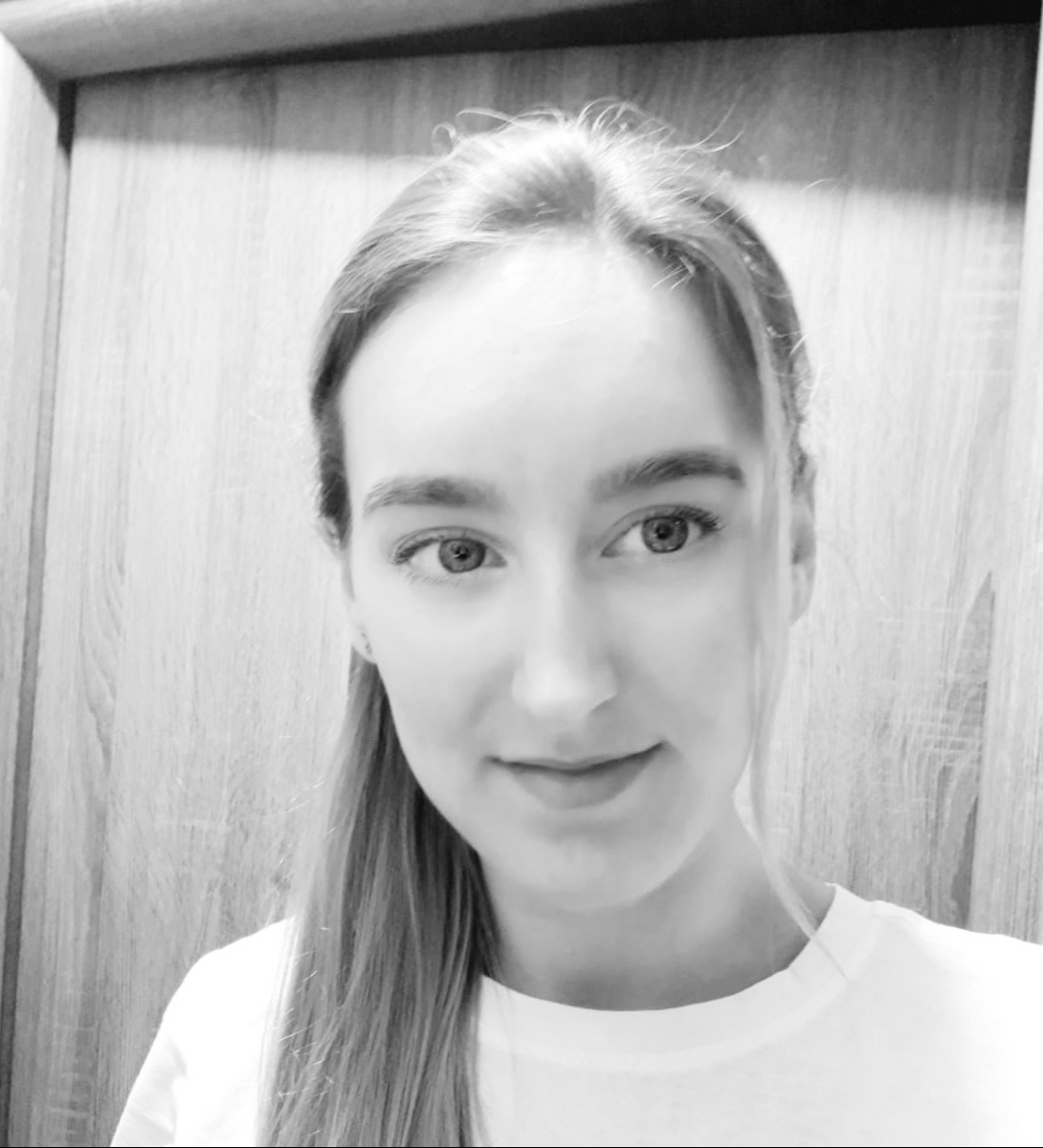 Czarno-białe portretowe zdjęcie młodej kobiety z włosami związanymi w kucyk, uśmiechającej się delikatnie do kamery na tle drewnianej ściany.