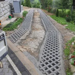 Dwa pasy ażurowych płyt betonowych, częściowo ułożonych w gruncie, prowadzące w kierunku wzgórz zalesionych w tle, widok z perspektywy niskiego kąta.