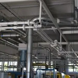 Złożony system rur metalowych, prawdopodobnie wentylacyjnych lub przemysłowych, w hali produkcyjnej w Ciechanowie. Widoczne liczne złączenia i zawory. Całość konstrukcji podwieszona pod sufitem.