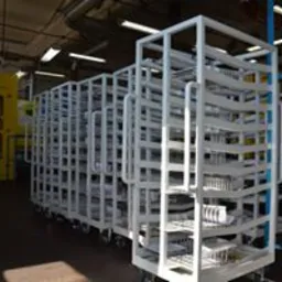 Metalowy w&oacute;zek transportowy z białymi ramami, wypełniony elementami metalowymi w hali produkcyjnej, oświetlenie g&oacute;rne.