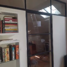 Metalowa konstrukcja z szybą, oddzielająca pomieszczenia, widoczna drabina w tle i regał z książkami po lewej stronie