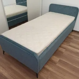 Niebieskie łóżko tapicerowane z pikowanym zagłówkiem i materacem, widoczne na drewnianej podłodze, z odbiciem w szafie z lustrem.