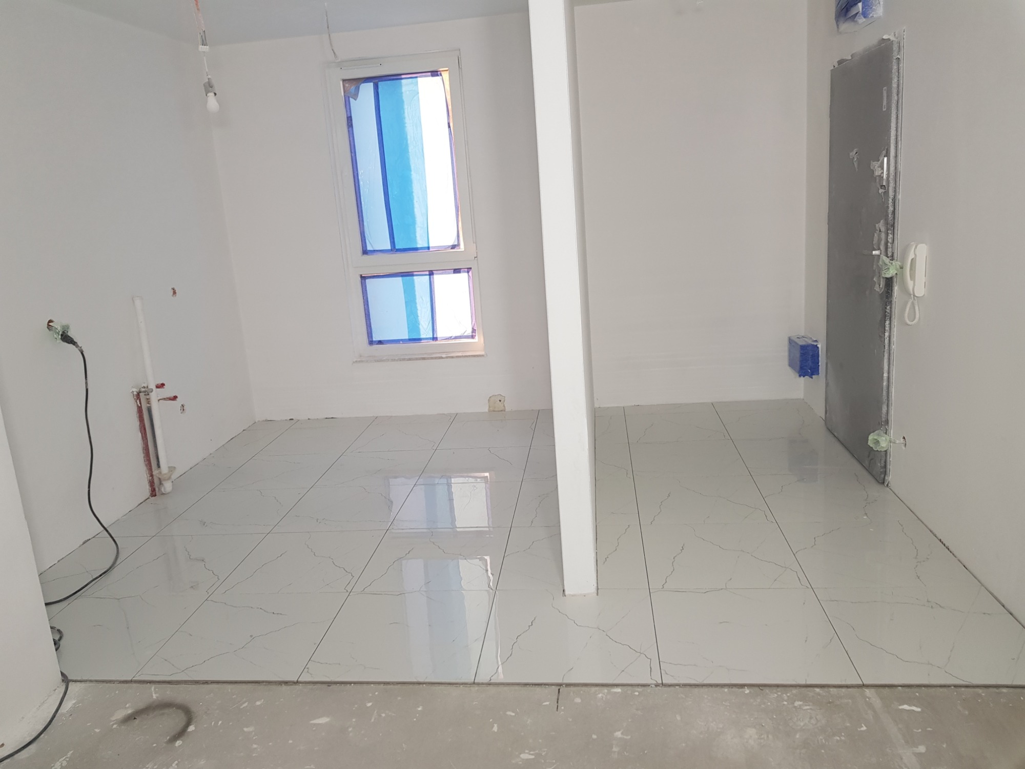 Wnętrze pokoju w trakcie układania białych płytek podłogowych z szarym wzorem żyłek, zabezpieczone okno, widoczne instalacje hydrauliczne i elektryczne.