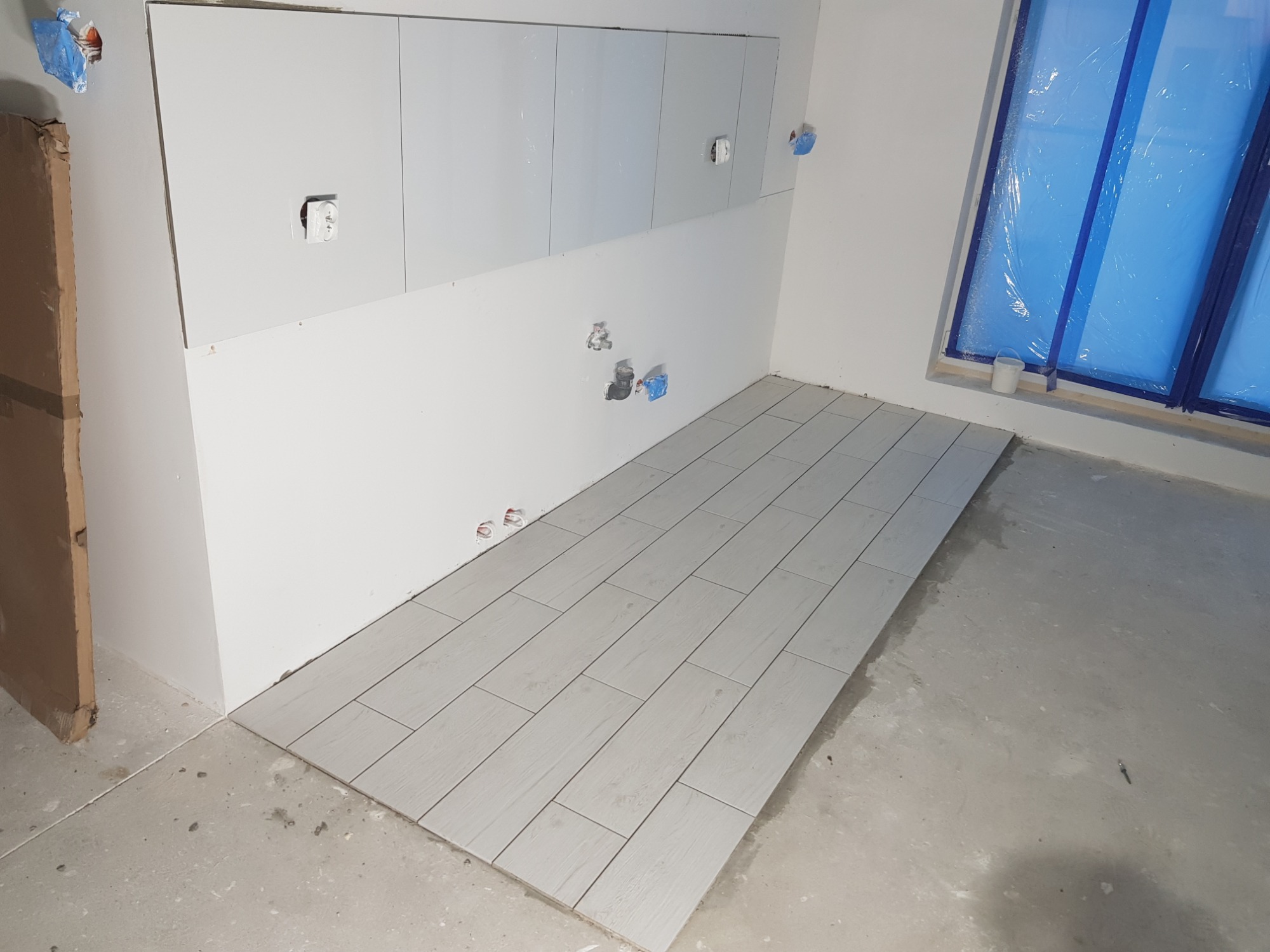 Układanie jasnoszarych płytek podłogowych w trakcie remontu łazienki, widoczne przygotowane przyłącza wodne i zabezpieczone folią okno.