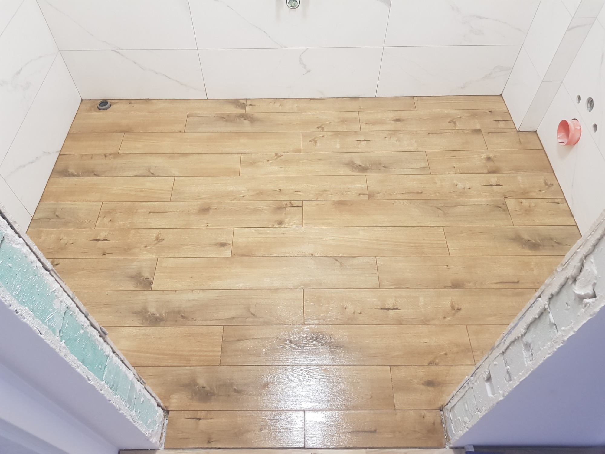 Podłoga imitująca drewno w kabinie prysznicowej z białymi płytkami na ścianach i widocznymi elementami instalacji.