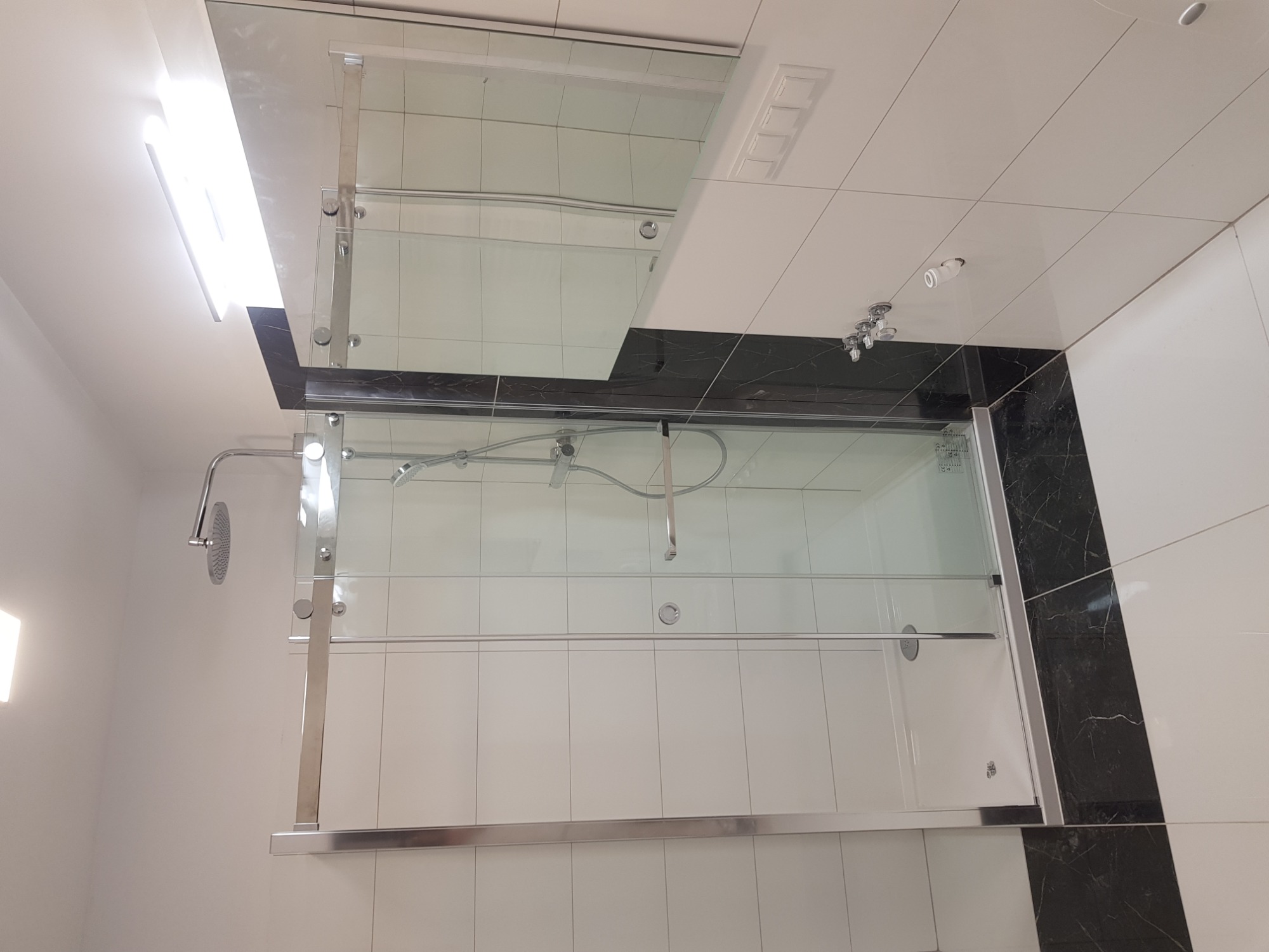 Kabina prysznicowa z przezroczystego szkła z chromowanymi okuciami, zamontowana w łazience z białymi i czarnymi płytkami. Widoczna deszczownica i bateria prysznicowa.