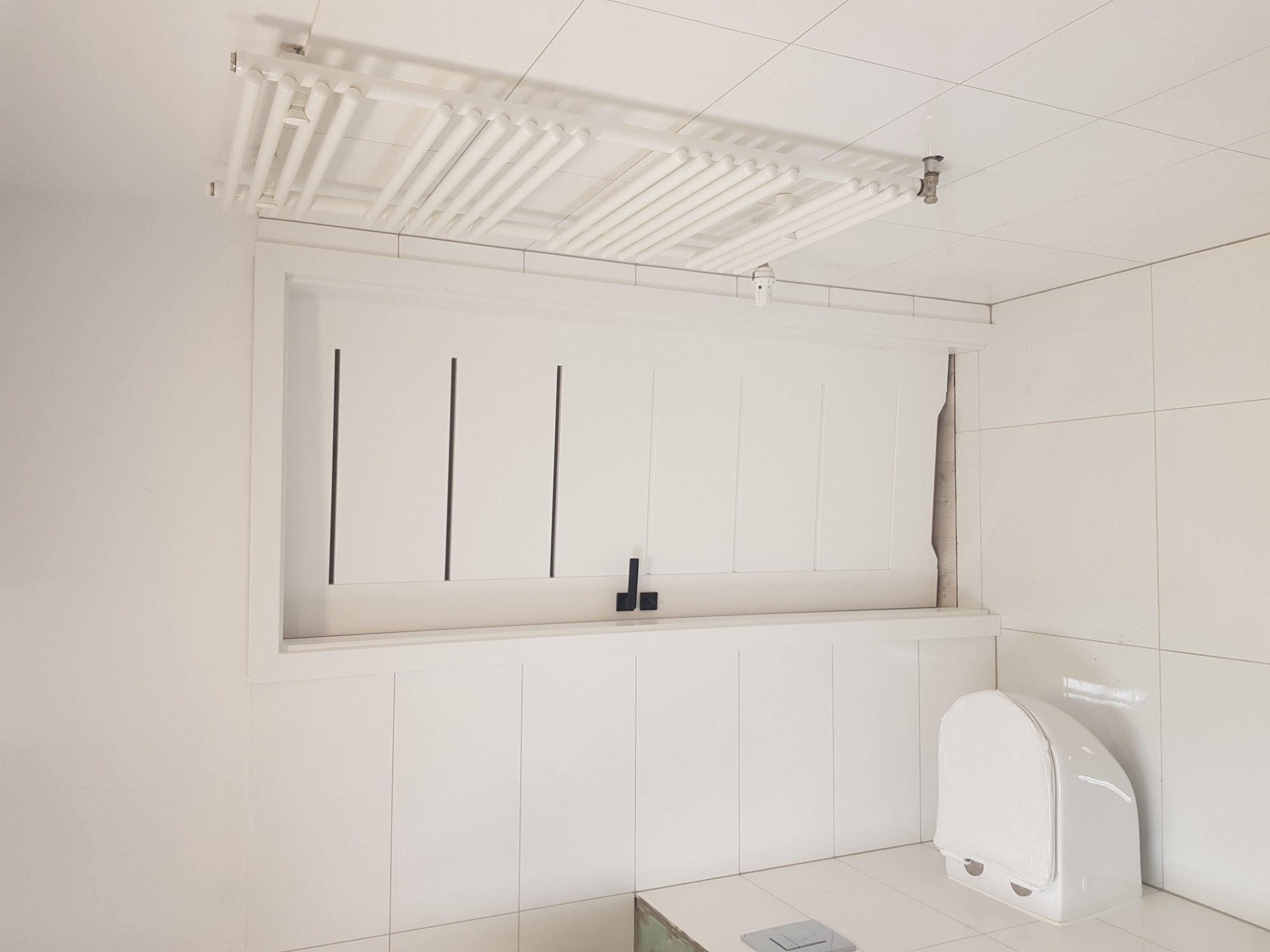 Biała łazienka z widocznymi rurami pod sufitem, białą zabudową ściany z pionowymi czarnymi paskami i toaletą ze specjalnym pokrowcem.