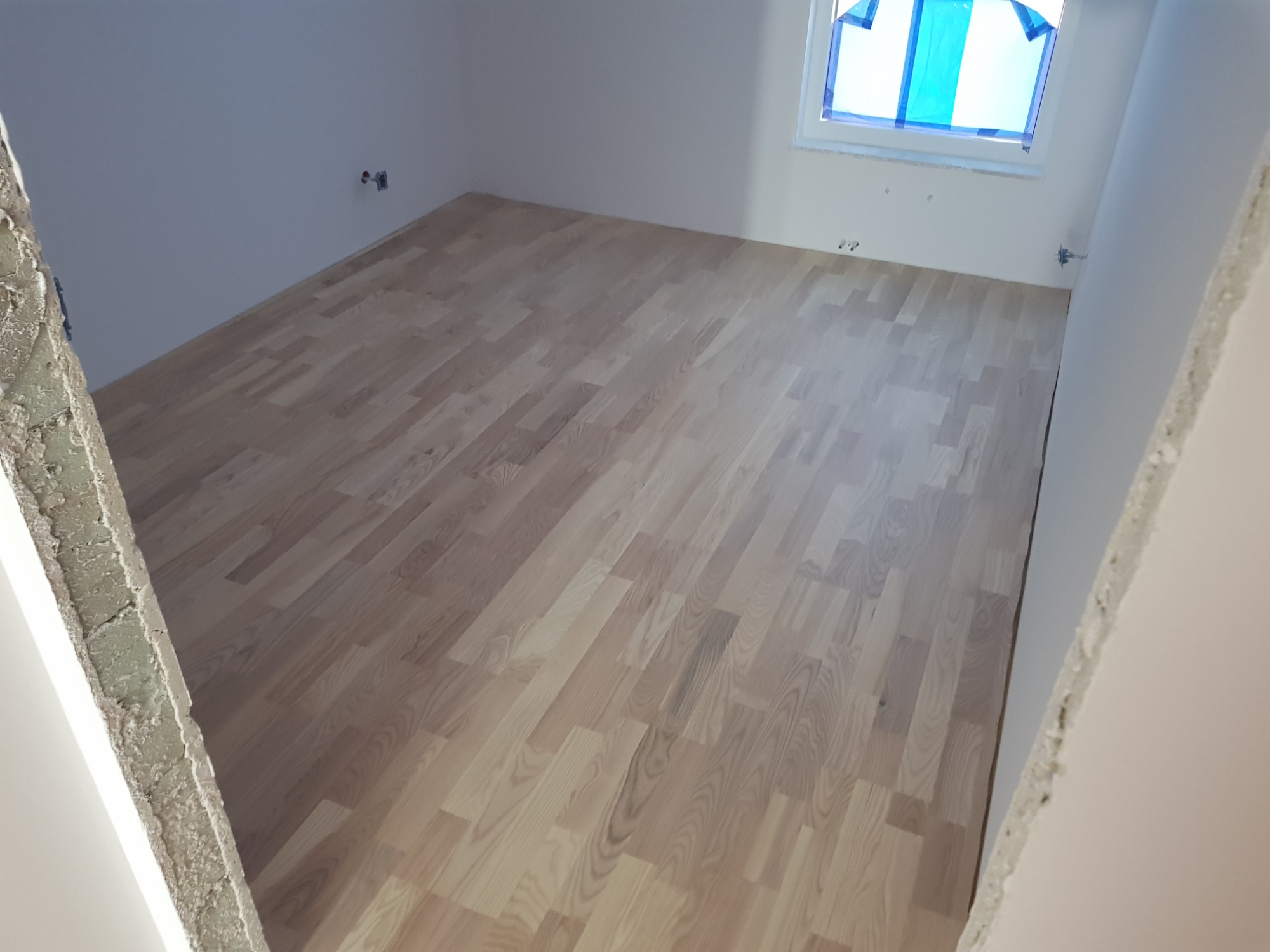 Jasny parkiet ułożony w pustym pokoju z oknem zabezpieczonym folią malarską, widok z perspektywy progu z surowym wykończeniem ściany.