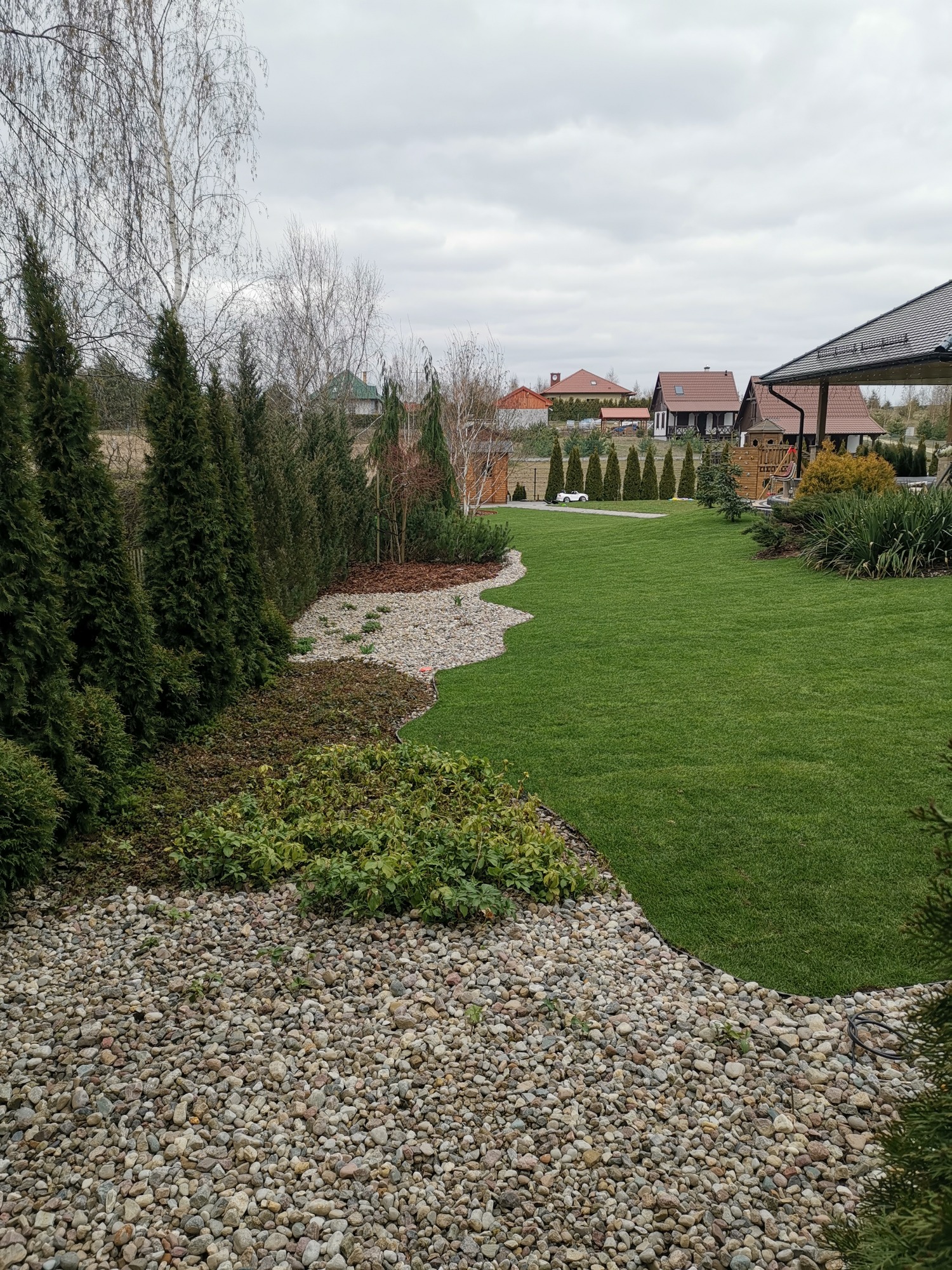 Starannie zaprojektowany ogród z równo przystrzyżonym trawnikiem, ozdobnymi rabatami wysypanymi kamykami i korą, oraz rzędem wysokich tuj w tle, tworzący uporządkowaną przestrzeń.