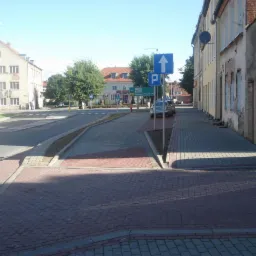 Zatoka postojowa w miejskiej przestrzeni, wyłożona czerwoną kostką brukową z szarymi obrzeżami, widoczny znak drogowy wskazujący kierunek jazdy i miejsce parkingowe, w tle budynki mieszkalne i drzewa.