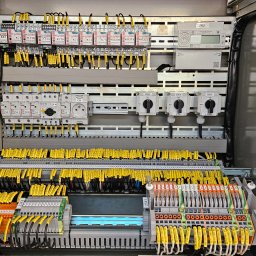 inst-el Kamil Łukasik - Rozdzielnica elektryczna z licznymi przewodami oznaczonymi żółtymi etykietami, widoczne bezpieczniki, przełączniki i inne elementy instalacji w szarej obudowie.