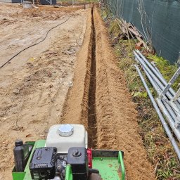 inst-el Kamil Łukasik - Wąski wykop w ziemi, wykonany przy użyciu specjalistycznej maszyny. W tle widoczny dom w trakcie budowy i ogrodzenie z siatki.