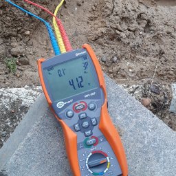 inst-el Kamil Łukasik - Miernik Sonel MPI-507 z podłączonymi przewodami pomiarowymi, leżący na betonowym bloku, w tle ziemia. Wyświetlacz pokazuje wynik pomiaru rezystancji uziemienia.