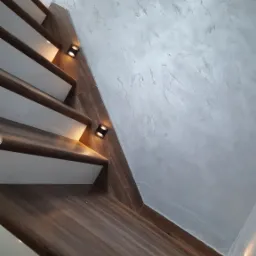Schody z drewnianymi stopniami i białymi podstopnicami, oświetlone małymi, kwadratowymi lampkami LED w ścianie, z dekoracyjną, szarą fakturą na ścianie i suficie.