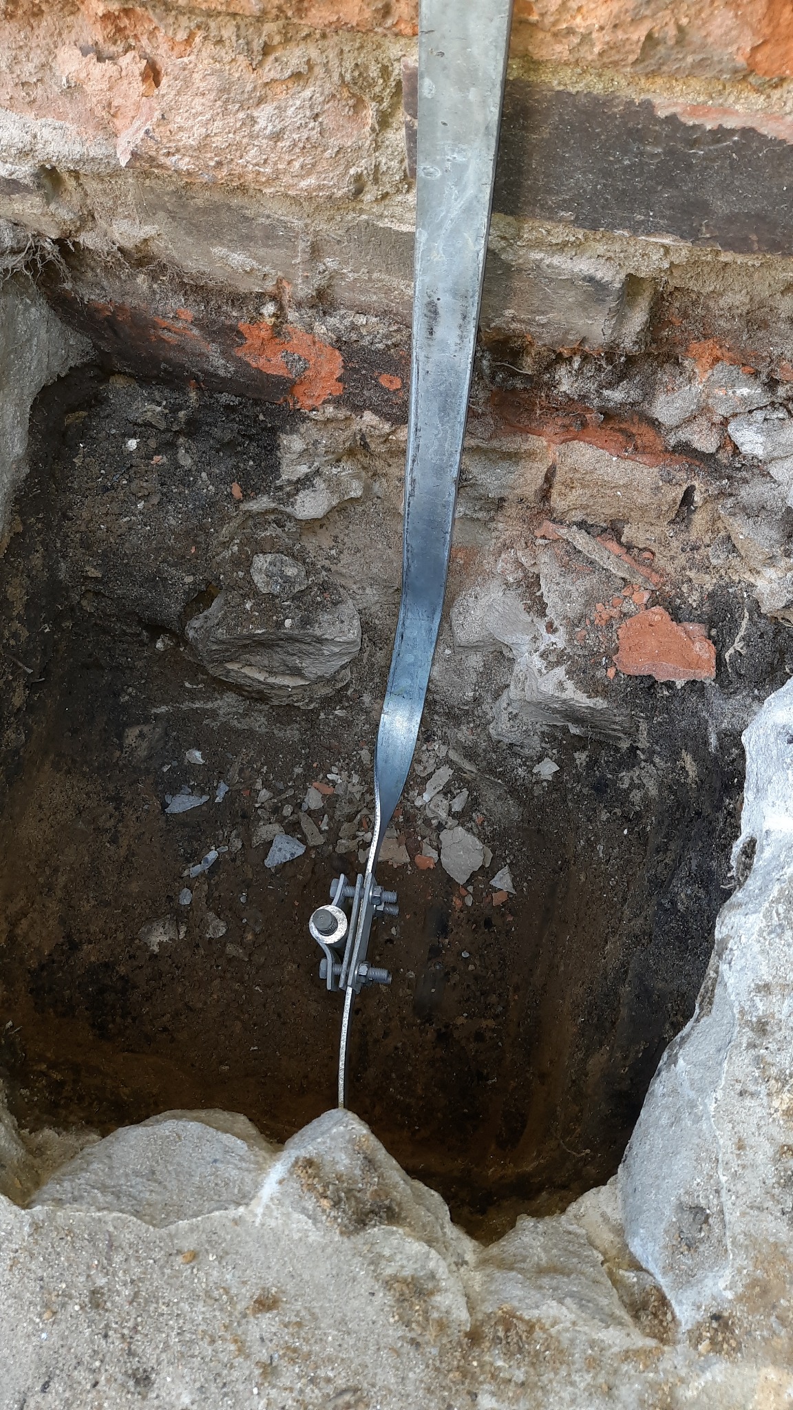 Metalowa listwa uziemiająca, przymocowana śrubami do przewodu w wykopie, na tle ceglanej ściany. Widoczne elementy instalacji odgromowej w ziemi.