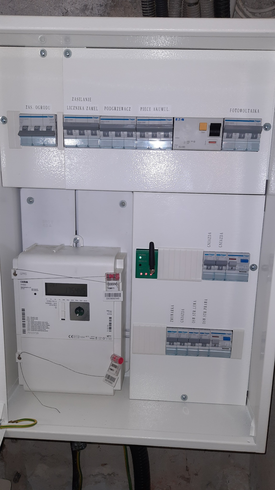 Rozdzielnia elektryczna w białej obudowie, z widocznym licznikiem energii, bezpiecznikami i opisami obwodów, w tym fotowoltaiki. Widoczne przewody i elementy montażowe.