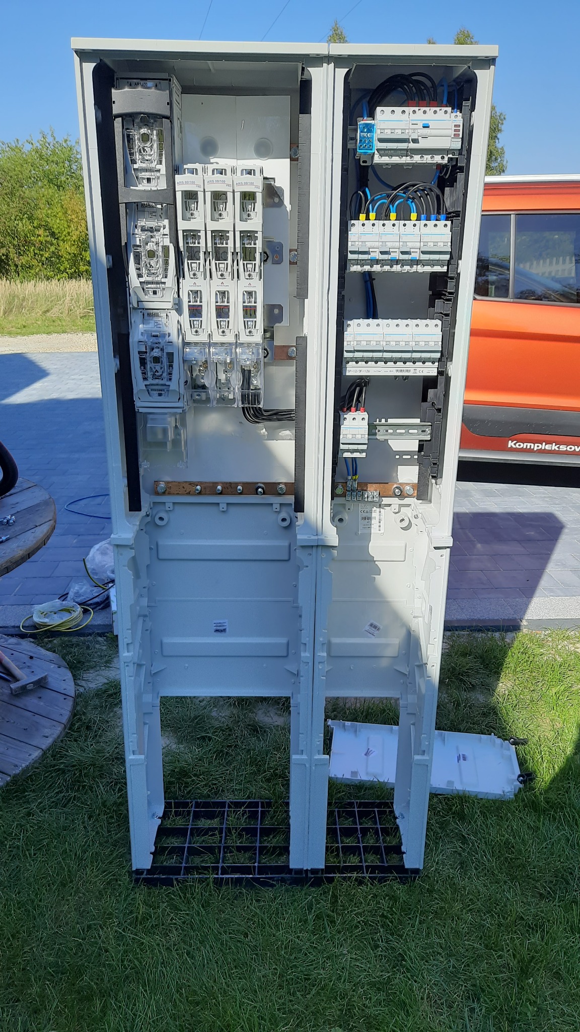 Otwarta, biała skrzynka elektryczna z zainstalowanymi licznikami i bezpiecznikami, widoczna na zewnątrz budynku. W tle fragment pomarańczowego samochodu.