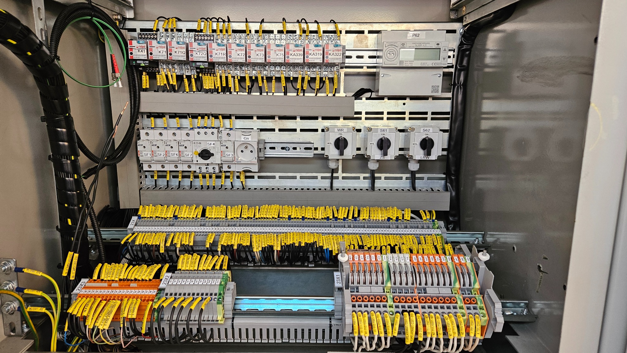 Rozdzielnica elektryczna z licznymi przewodami oznaczonymi żółtymi etykietami, widoczne bezpieczniki, przełączniki i inne elementy instalacji w szarej obudowie.