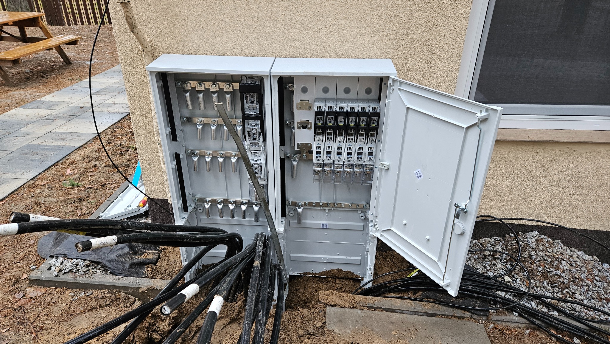 Otwarta, biała skrzynka elektryczna z licznymi kablami na zewnątrz, umieszczona na ziemi przy ścianie budynku z oknem. Widoczne elementy instalacji wewnątrz.