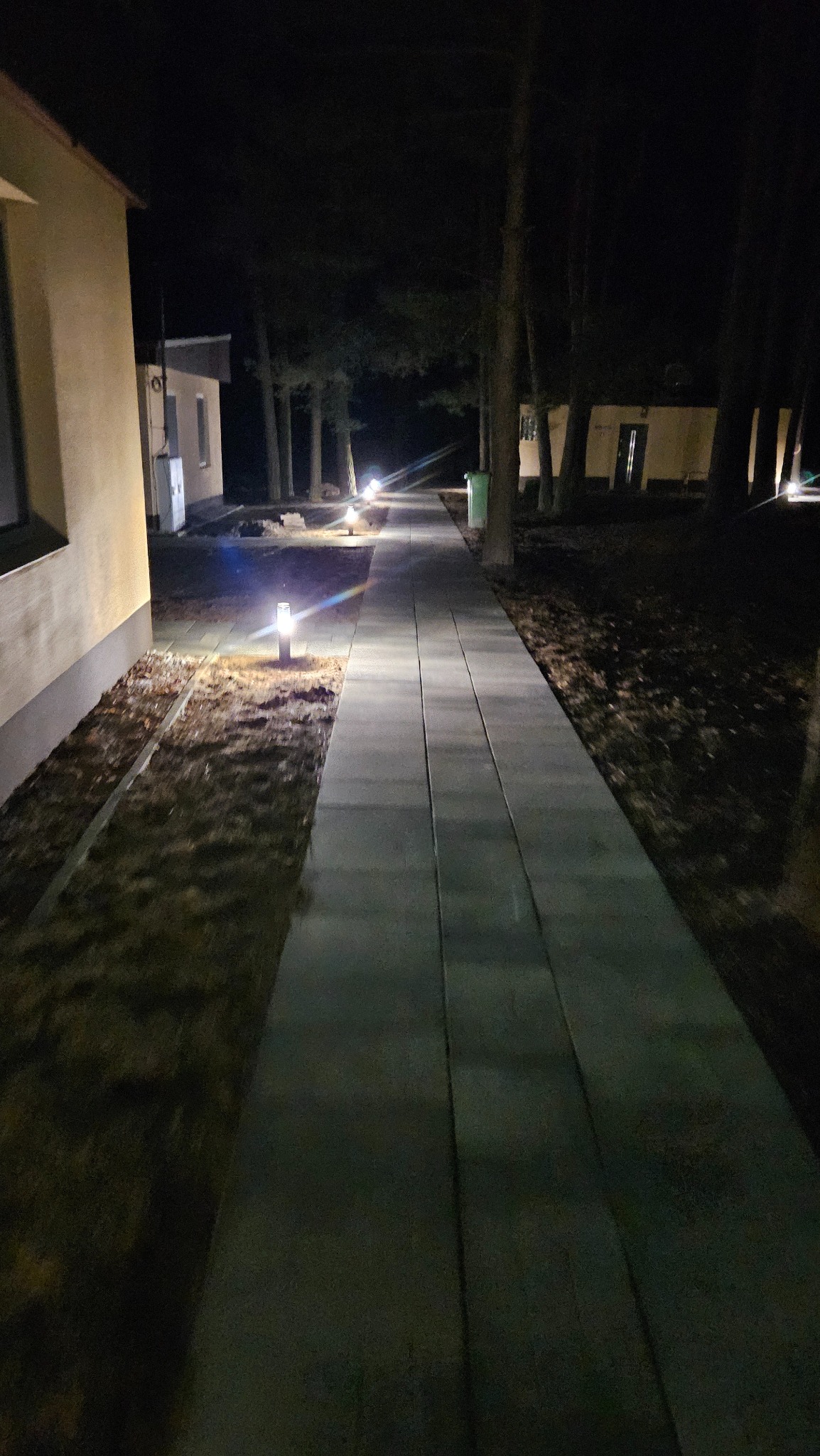 Oświetlona nocą ścieżka z kostki brukowej prowadząca wzdłuż budynku, z lampami ogrodowymi po bokach, oświetlającymi drogę w ciemności.