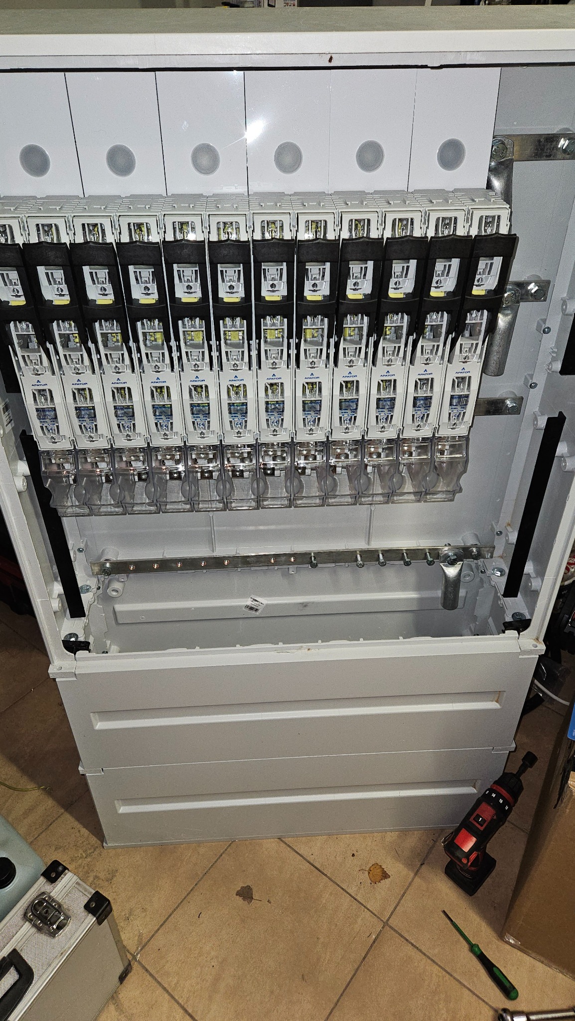 Rozdzielnica elektryczna w trakcie montażu, widoczne zabezpieczenia i szyny montażowe. Na podłodze narzędzia: wkrętarka i śrubokręt.