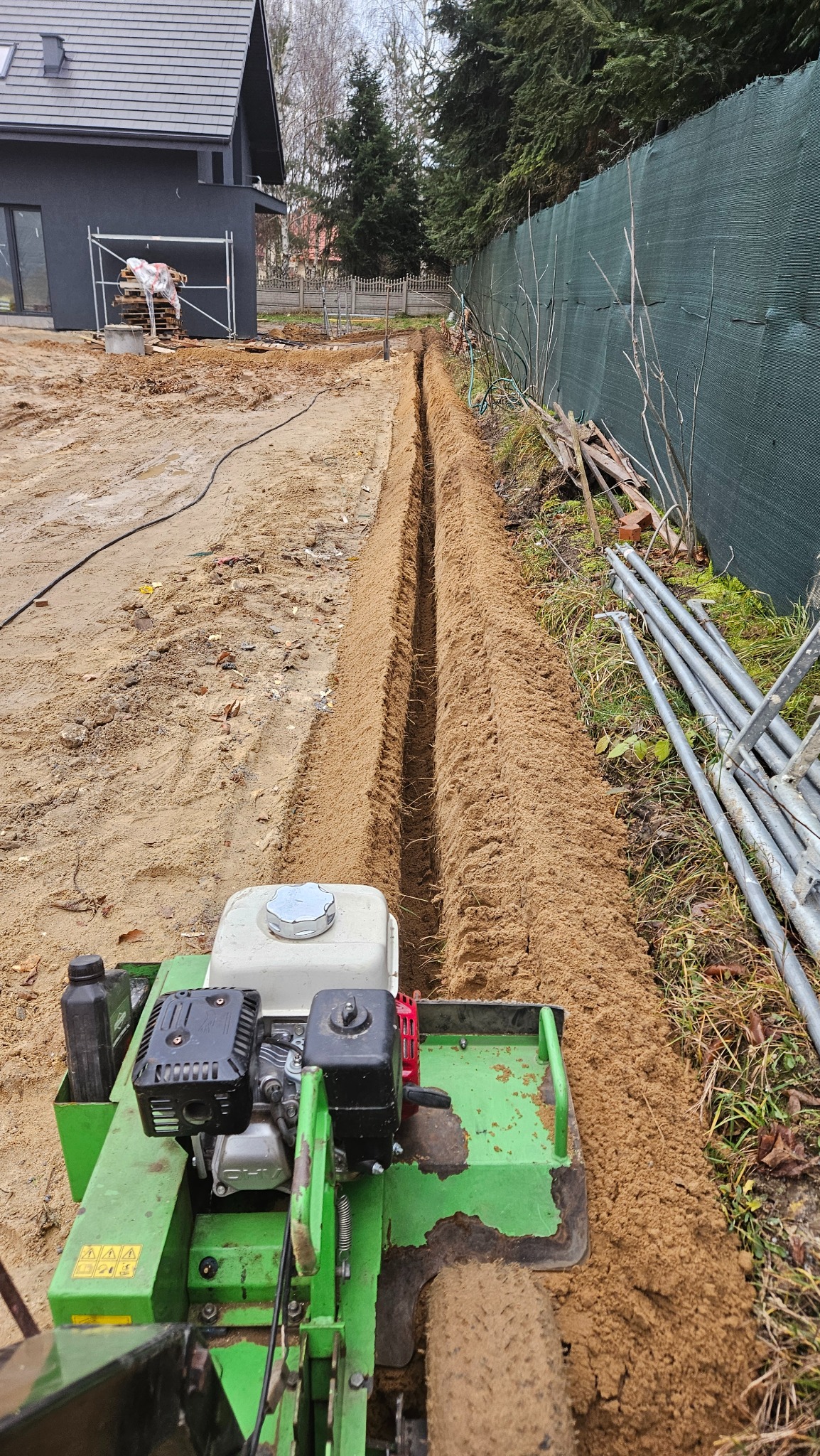 Wąski wykop w ziemi, wykonany przy użyciu specjalistycznej maszyny. W tle widoczny dom w trakcie budowy i ogrodzenie z siatki.