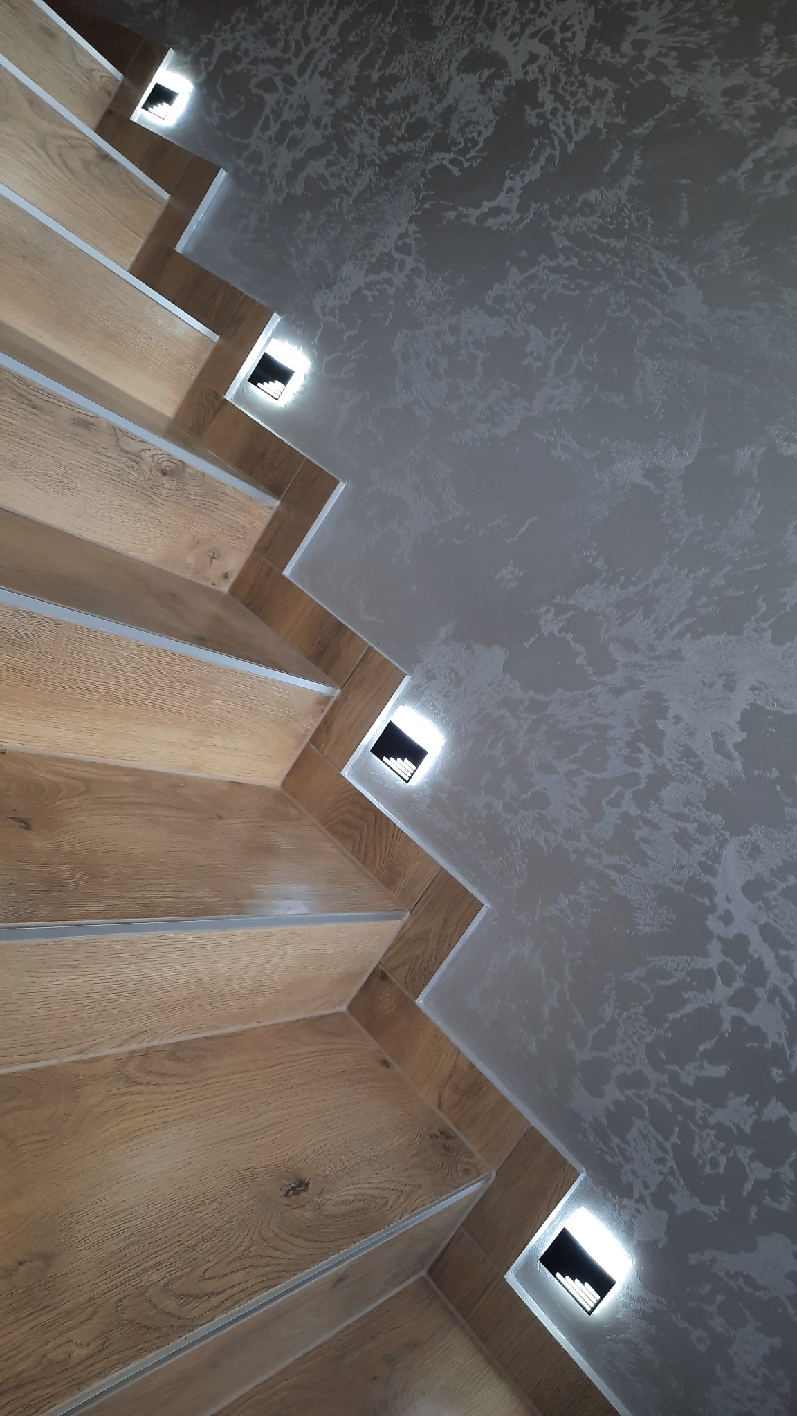 Drewniane schody z podświetleniem LED w ścianie, widok z dołu. Dekoracyjna ściana w tle. Nietypowy kąt ujęcia podkreśla nowoczesny design.