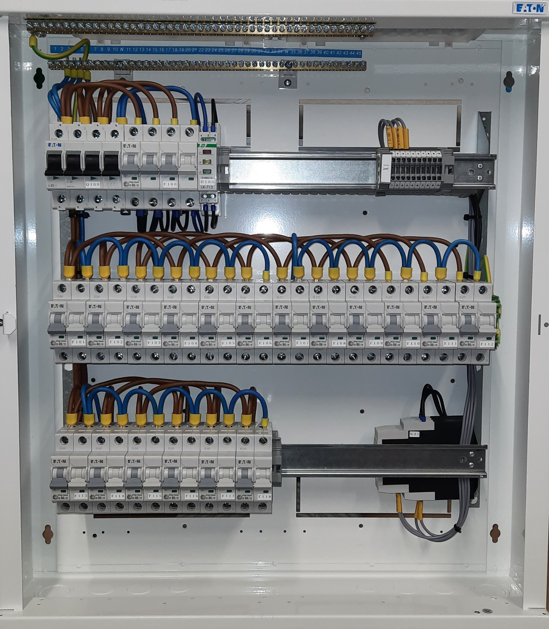 Rozdzielnica elektryczna marki Eaton z licznymi bezpiecznikami i precyzyjnie ułożonymi przewodami w kolorach niebieskim, brązowym i żółto-zielonym, zamontowana w białej obudowie.