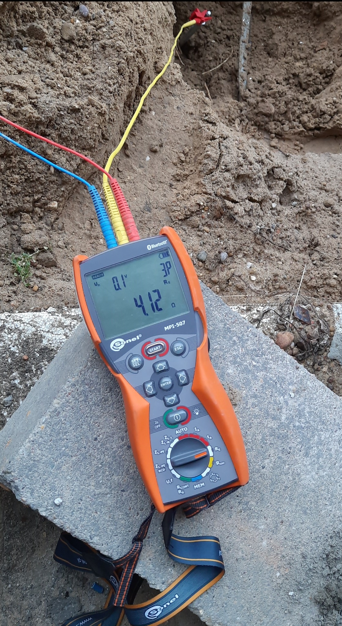 Miernik Sonel MPI-507 z podłączonymi przewodami pomiarowymi, leżący na betonowym bloku, w tle ziemia. Wyświetlacz pokazuje wynik pomiaru rezystancji uziemienia.