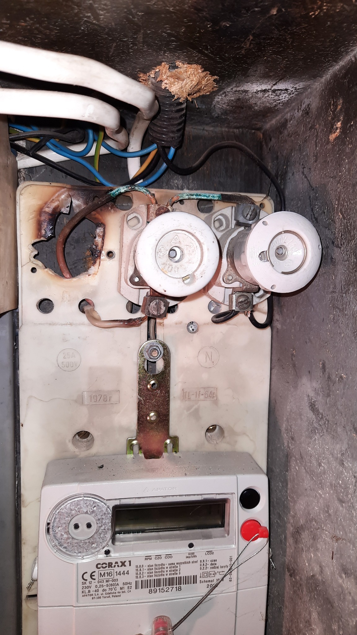 Stara skrzynka z bezpiecznikami topikowymi i licznikiem energii elektrycznej. Widoczne ślady przepalenia i uszkodzenia izolacji przewodów.