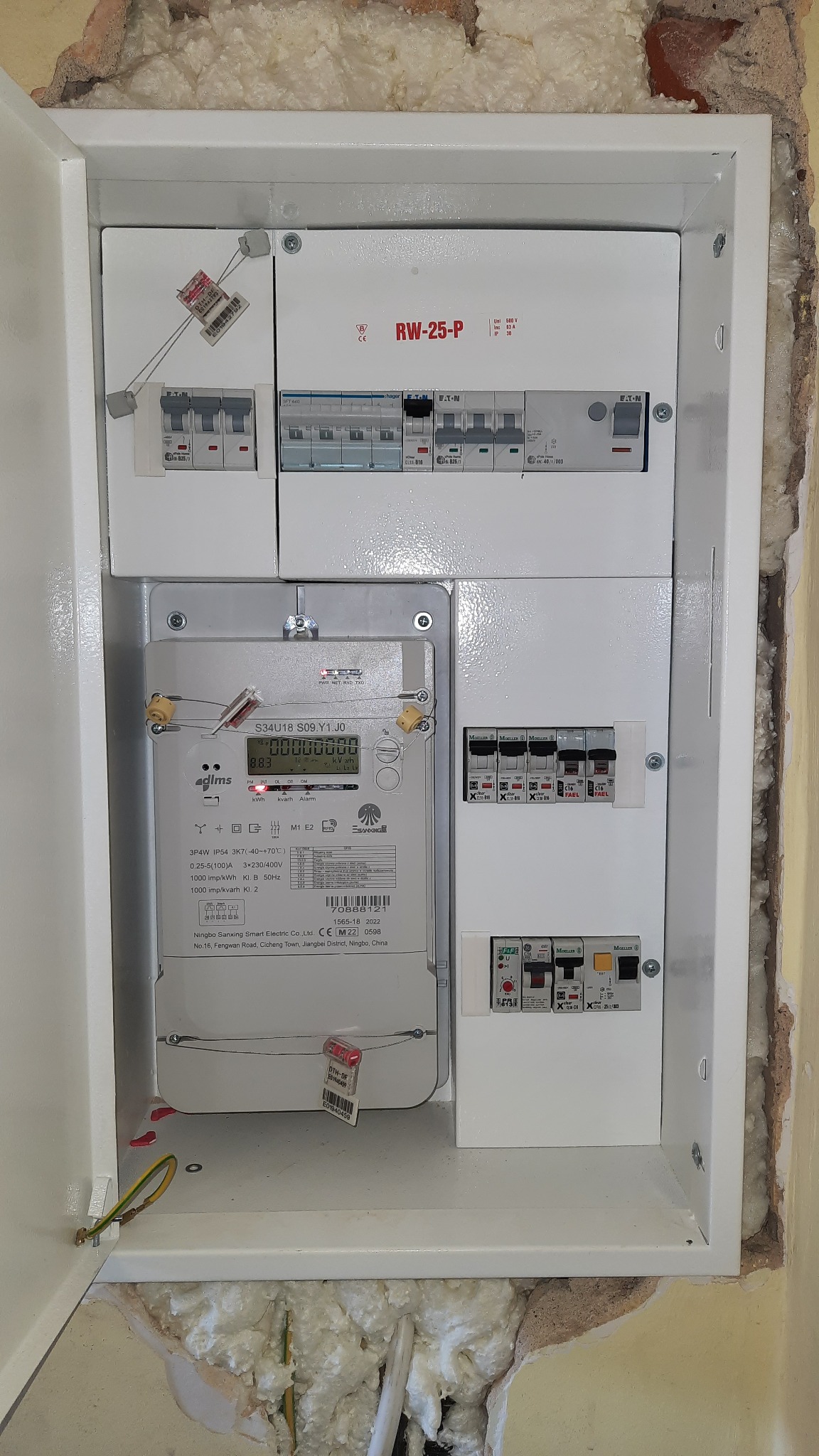 Nowa, biała skrzynka elektryczna z zamontowanym licznikiem i bezpiecznikami, widoczna instalacja w ścianie. Szczegółowe ujęcie na elementy instalacji elektrycznej.