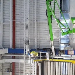 Monteraż opraw LED w hali przemysłowej z użyciem podnośnika koszowego; pracownik w kasku na wysokości mocuje oświetlenie do sufitu z widocznymi instalacjami.