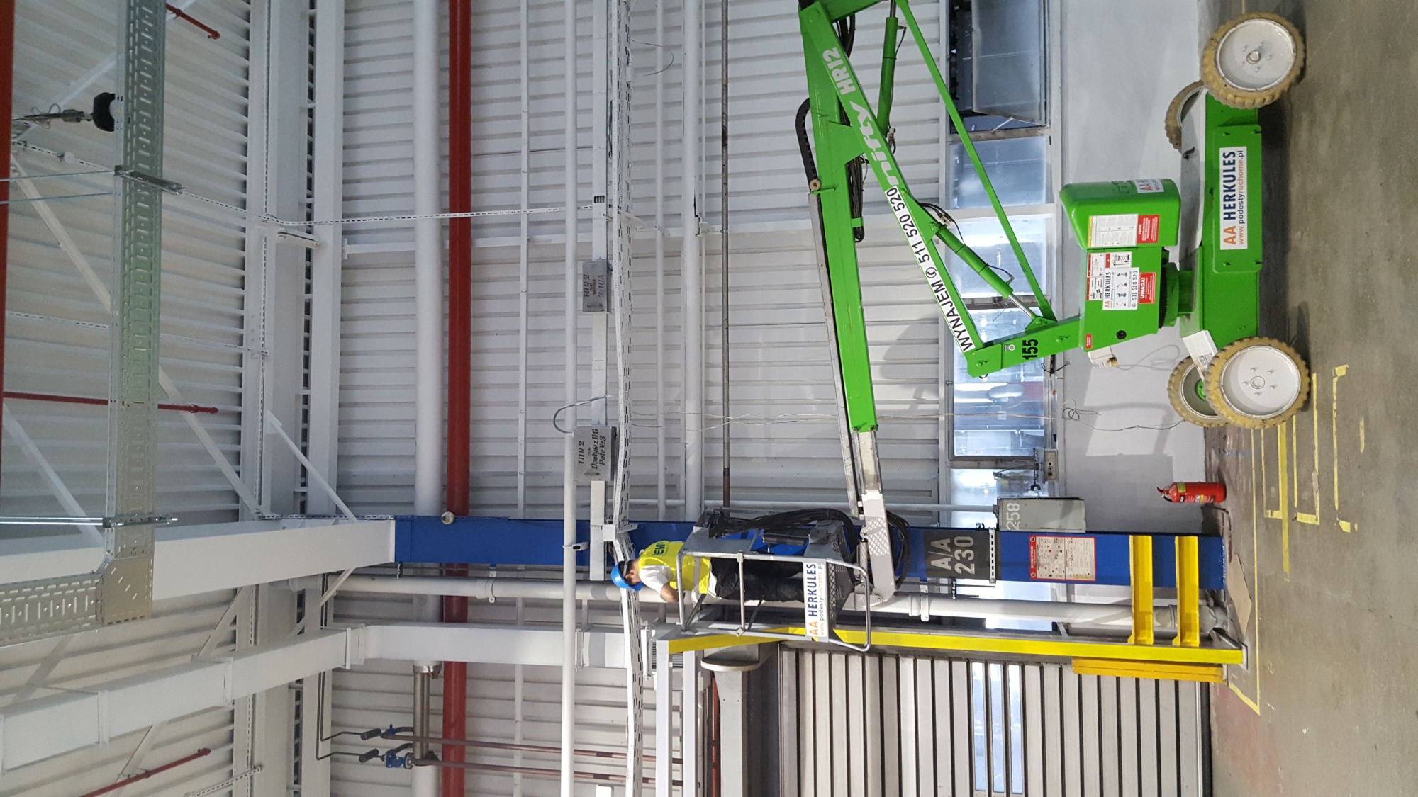 Monteraż opraw LED w hali przemysłowej z użyciem podnośnika koszowego; pracownik w kasku na wysokości mocuje oświetlenie do sufitu z widocznymi instalacjami.