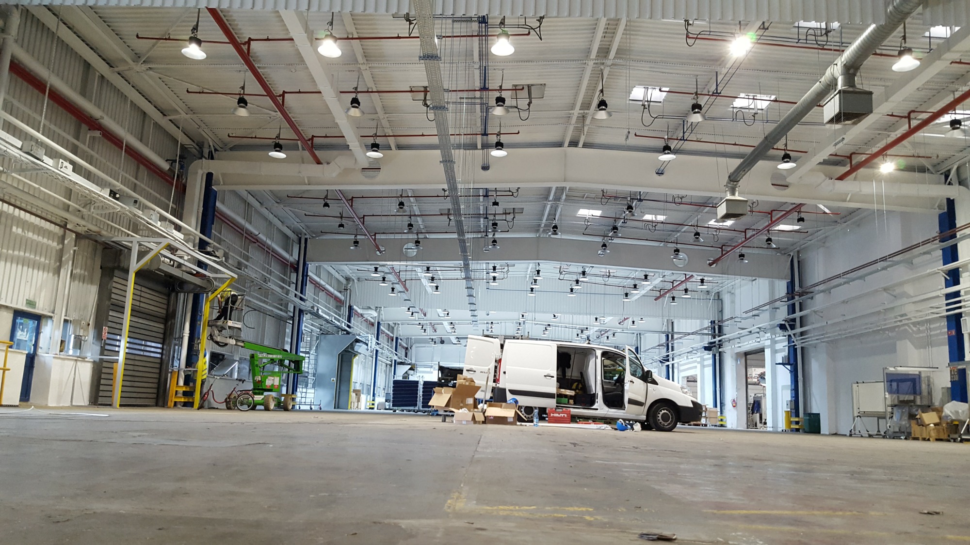 Montaż opraw LED w hali przemysłowej, widoczny podnośnik koszowy i samochód dostawczy z otwartymi drzwiami.